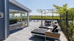 A modern outdoor patio with a glass wall, outdoor seating, and a view of a body of water.