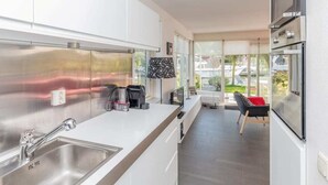 Una cocina moderna con electrodomésticos de acero inoxidable, una encimera blanca y vistas al exterior a través de amplios ventanales.