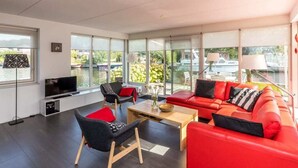 Una sala de estar moderna con un sofá rojo, sillas negras y rojas, un televisor y ventanas amplias con vistas a un cuerpo de agua.