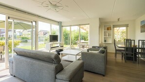 A spacious living room with a large TV, grey sofas, a dining area with chairs, and large windows offering a view of the garden and neighboring houses. - Beautiful home with roof terrace and a view of the water. (Oude-Tonge)