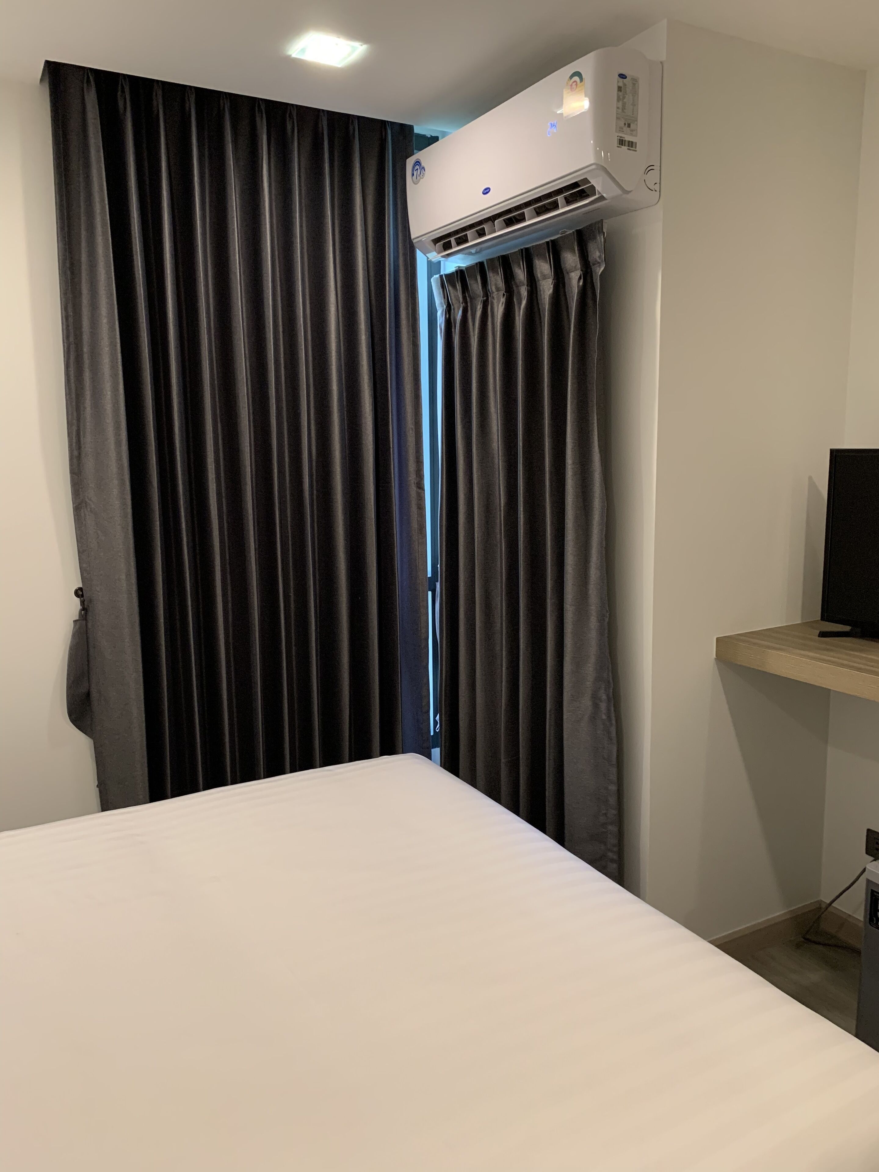 Quarto de hotel com cama, cortinas escuras, ar condicionado e televisão sobre uma prateleira de madeira.