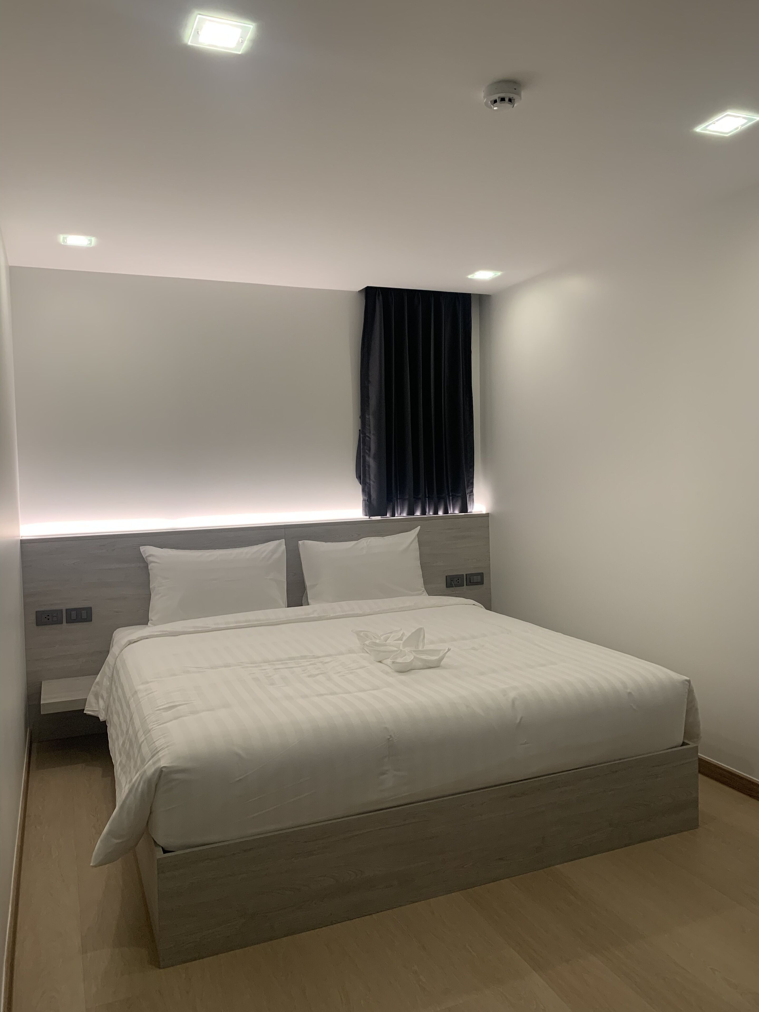 A modern hotel room with a large bed, a headboard with built-in lighting, a dark curtain, and a ceiling-mounted security camera.