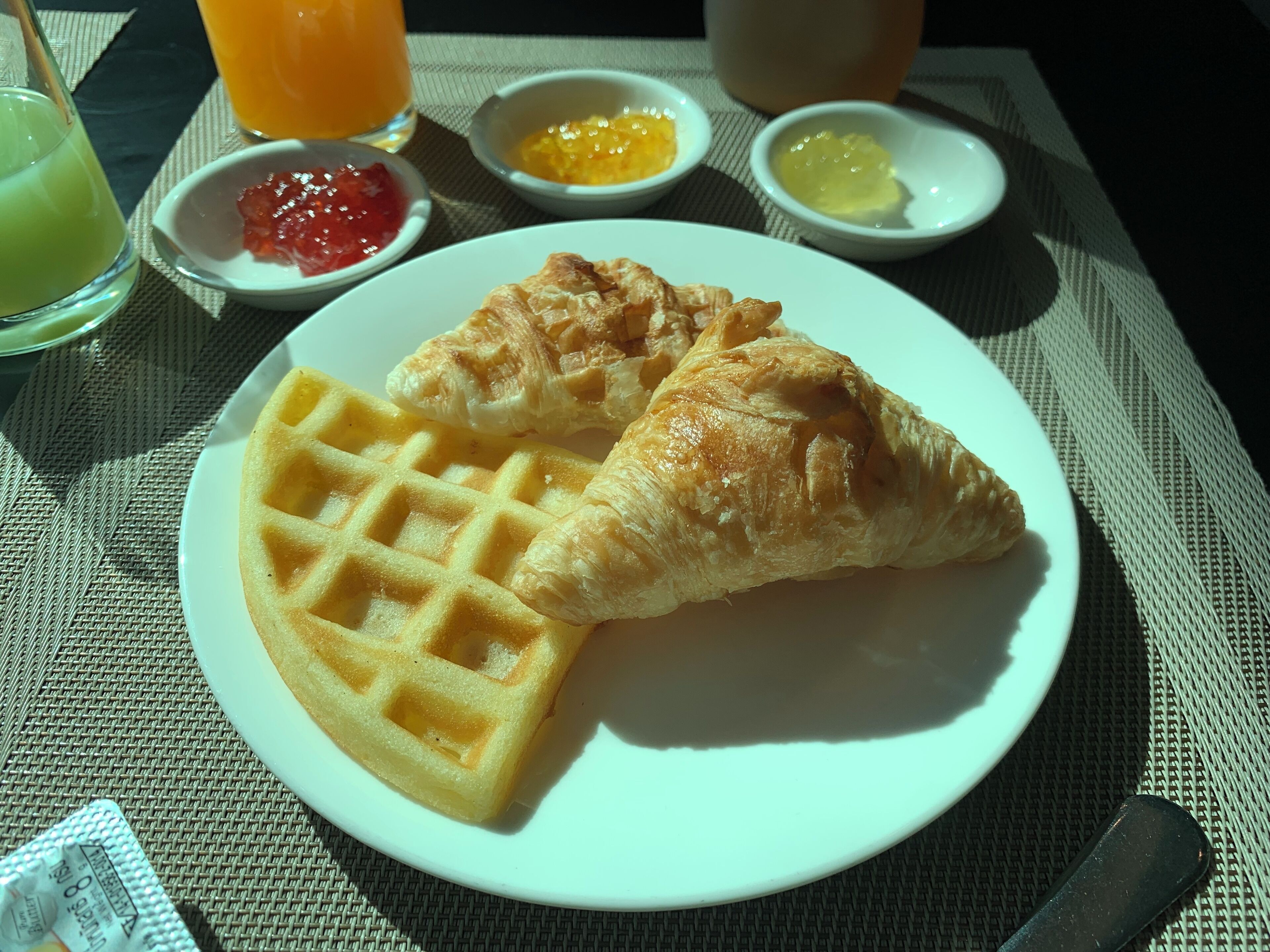 Ein Teller mit einem Waffel und Croissant, dazu kleine Schalen mit Marmelade und Butter, sowie ein Glas Orangensaft auf einem Tisch.