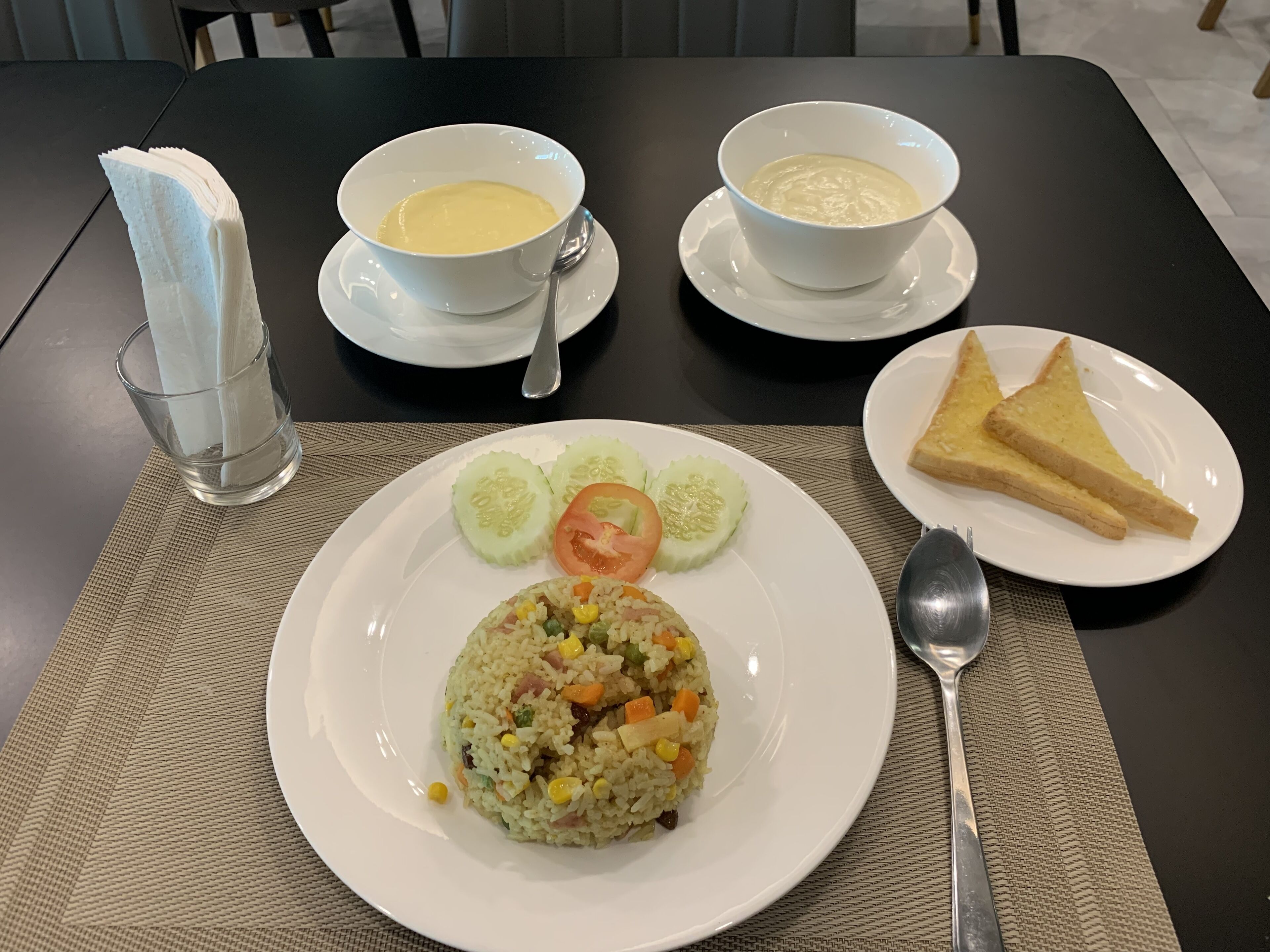 Une table avec une assiette de riz frit, un bol de soupe, un bol de yaourt, un verre d’eau et une assiette de toasts.