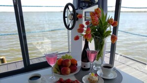 A table with a variety of food and drinks, including a bottle of wine, a glass of pink drink, and plates of fruit and pastries, set against a view of the water. - Domki na wodzie ARKA 1, rower wodny, do morza oraz centrum 400m (Mielno)