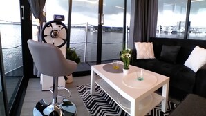 A modern interior with a sofa, a coffee table, a chair, and a view of the water through large windows. - Domki na wodzie ARKA 1, rower wodny, do morza oraz centrum 400m (Mielno)