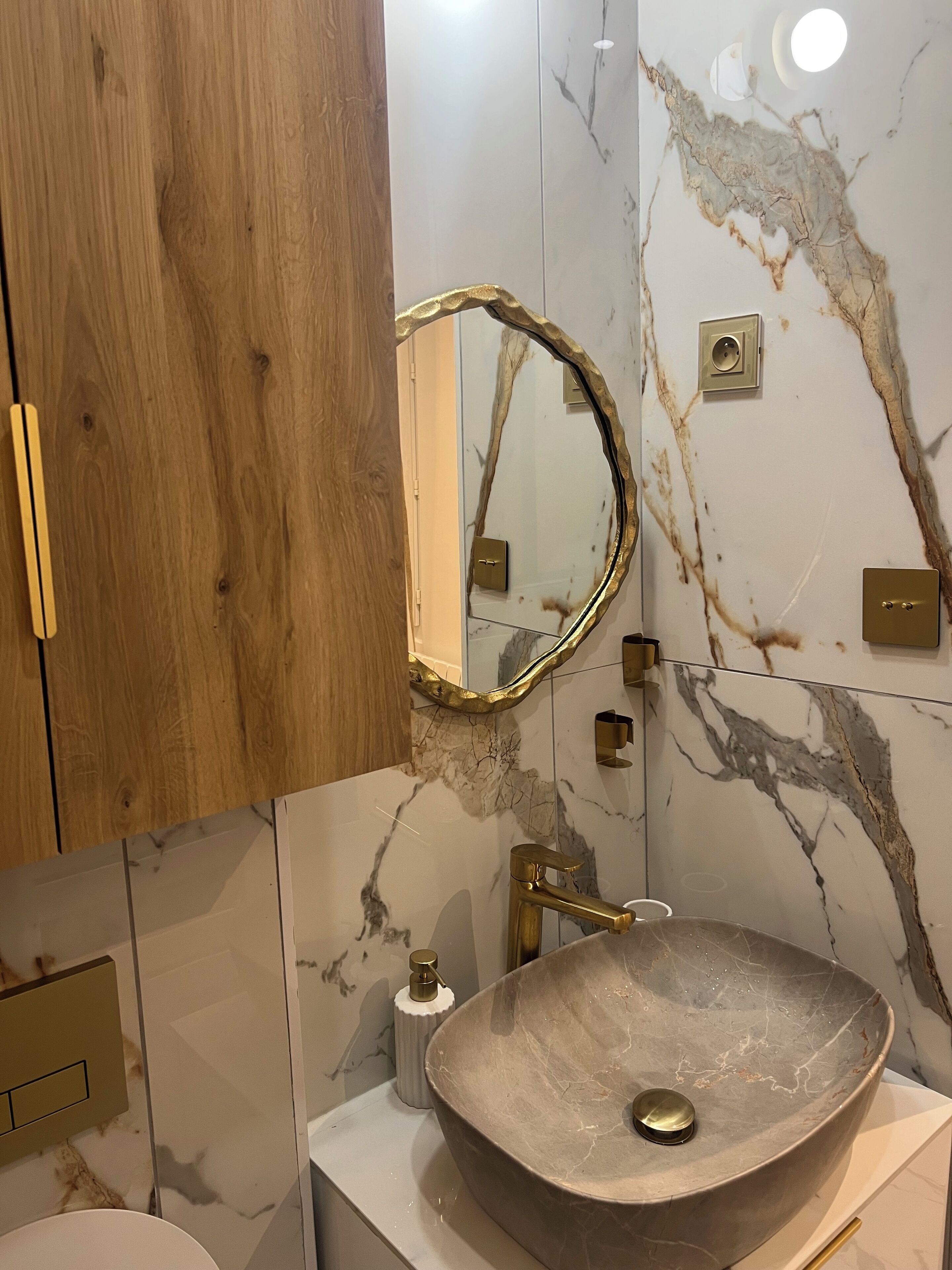 A bathroom with a wooden vanity, a marble backsplash, a gold-framed mirror, a vessel sink, and a gold faucet.