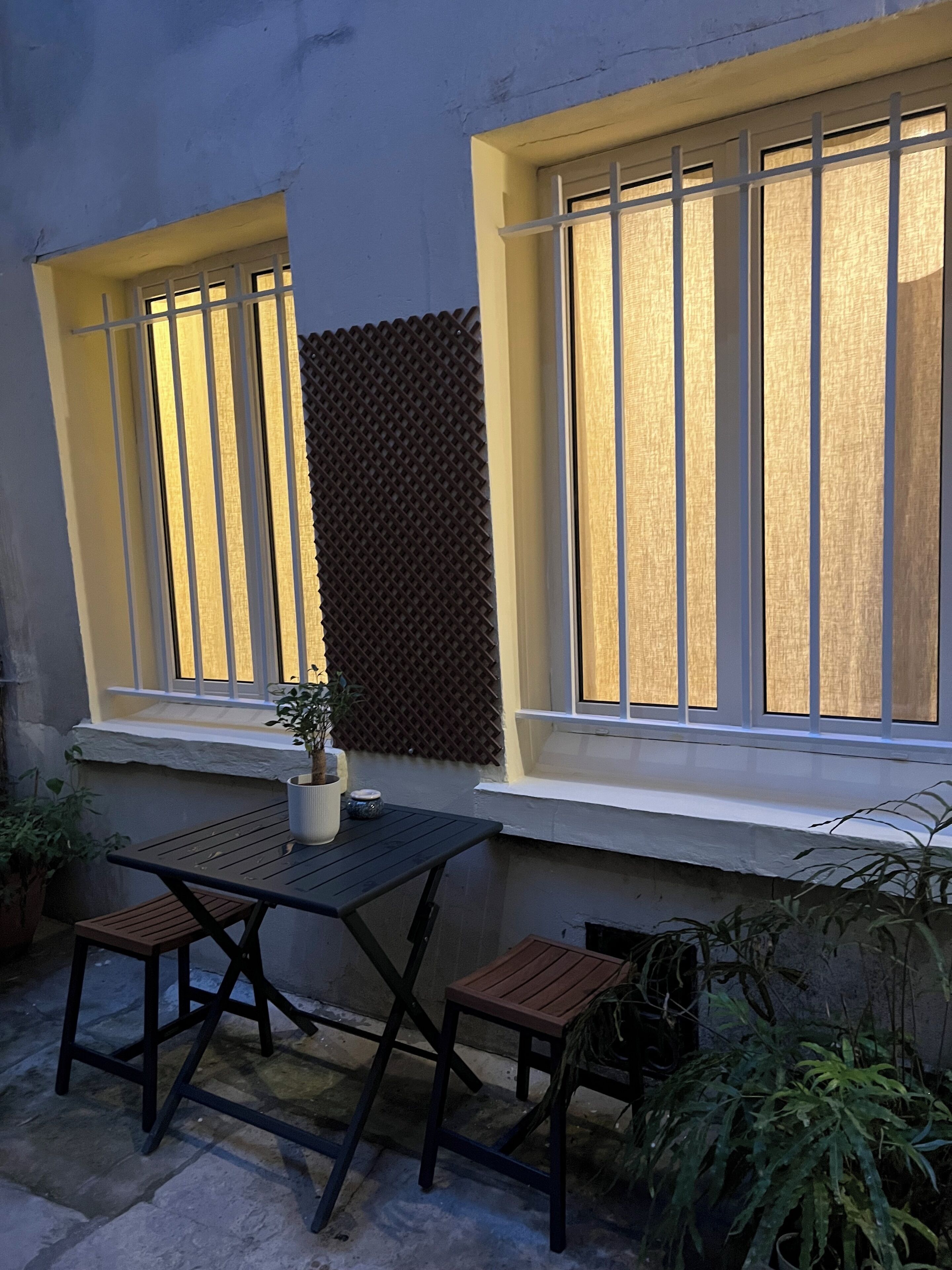 Une terrasse avec une table et des chaises, un pot de fleurs et une fenêtre dont le mur arrière présente une texture particulière.