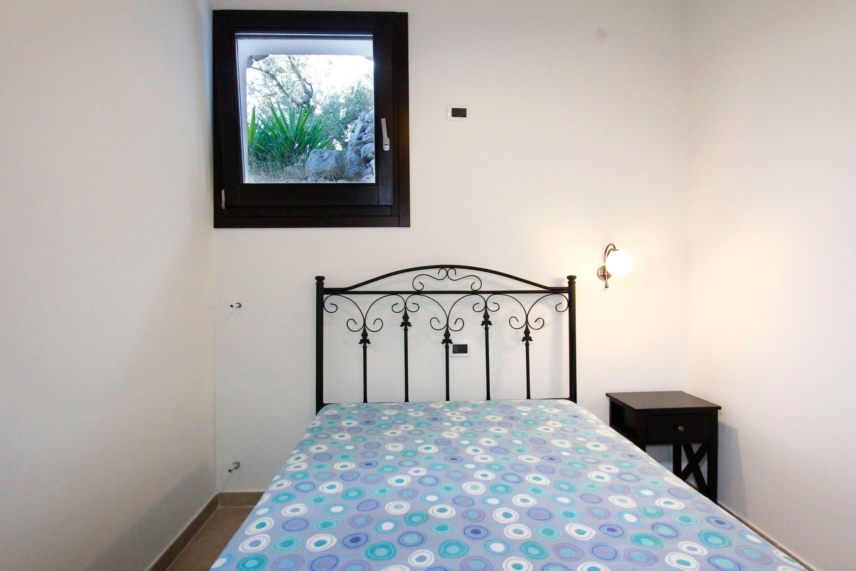 A bedroom with a bed, a nightstand, a wall-mounted light, and a window with a view of greenery.