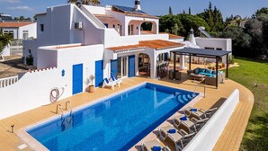 Una piscina con sillones de descanso y un área de estar techada, rodeada por un edificio blanco con detalles azules y un tejado de baldosas.