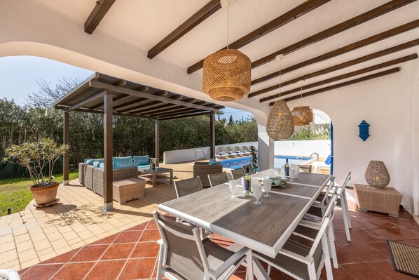 Un patio al aire libre con una mesa de madera, sillas y una zona de estar techada.