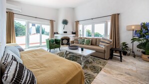 Un salón amplio con sofá, una mesa de centro y un sillón verde. Hay una chimenea con un abanico decorativo encima.