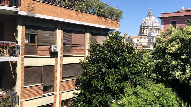 Un edifici de diversos pisos amb balcons, una gran estructura de cúpula al fons i vegetació exuberant al primer pla.