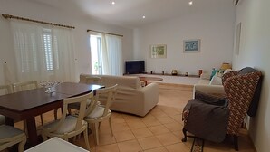 Uma sala de estar com uma mesa de jantar, cadeiras, um sofá, uma televisão e obras de arte nas paredes.