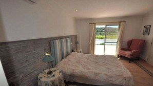 Quarto com cama, uma cadeira, mesinha de cabeceira e luminária, com vista para uma paisagem através da janela.