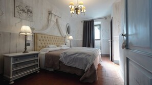 A bedroom with a tufted headboard, a bedside table with a lamp, a large bed with a blanket, and a chandelier.