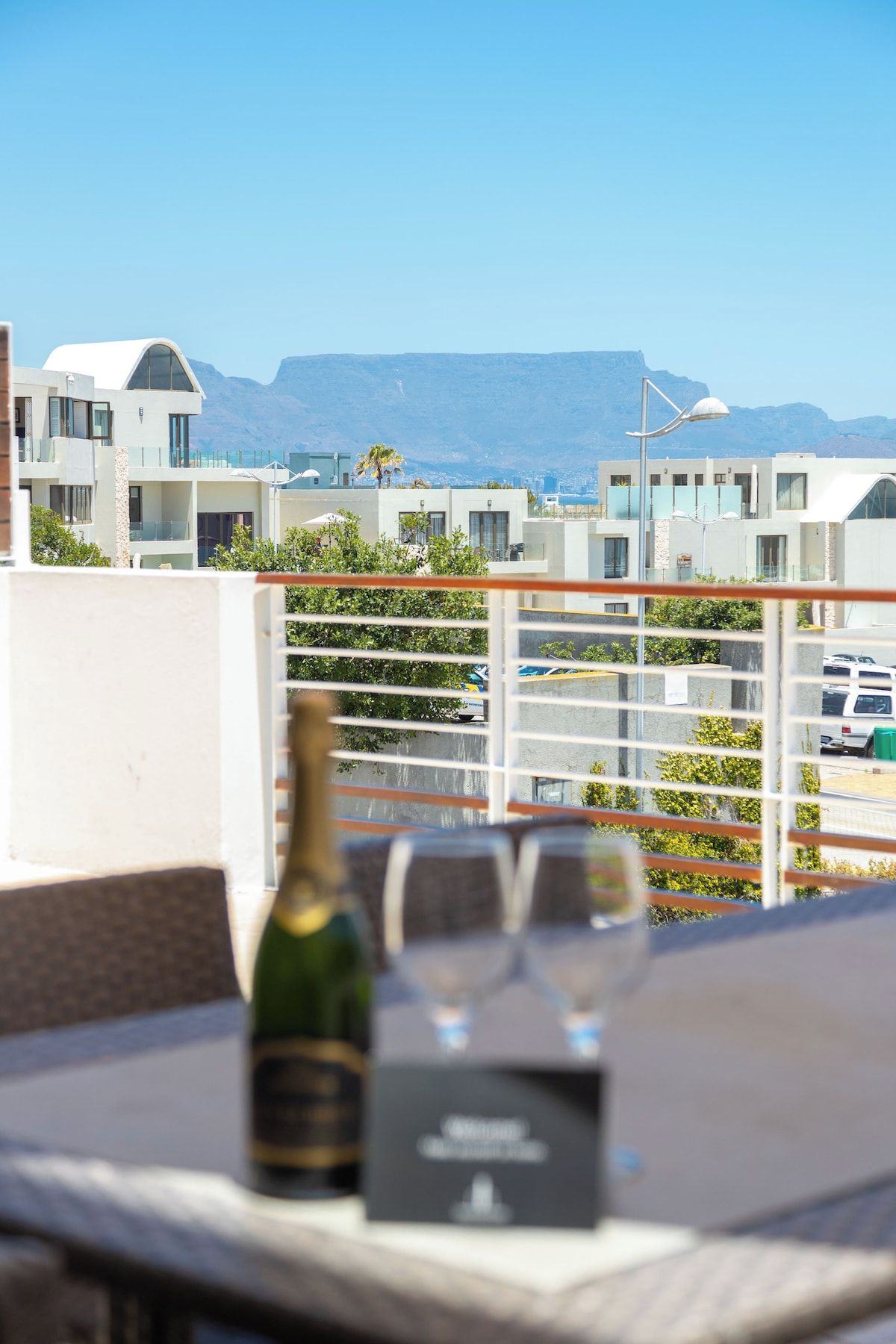 A balcony with a table, a bottle, and a glass, overlooking a cityscape with buildings and mountains.