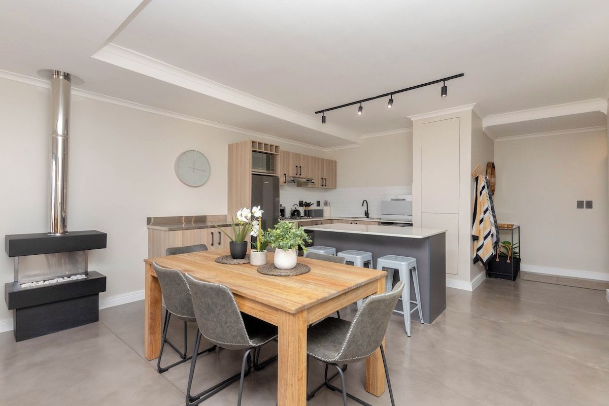 Une cuisine moderne avec un îlot central, une table à manger en bois et des chaises recouvertes d’un tissu gris.