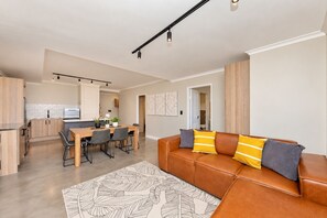 A modern living room with a leather sofa, a dining table with chairs, and a kitchen in the background.