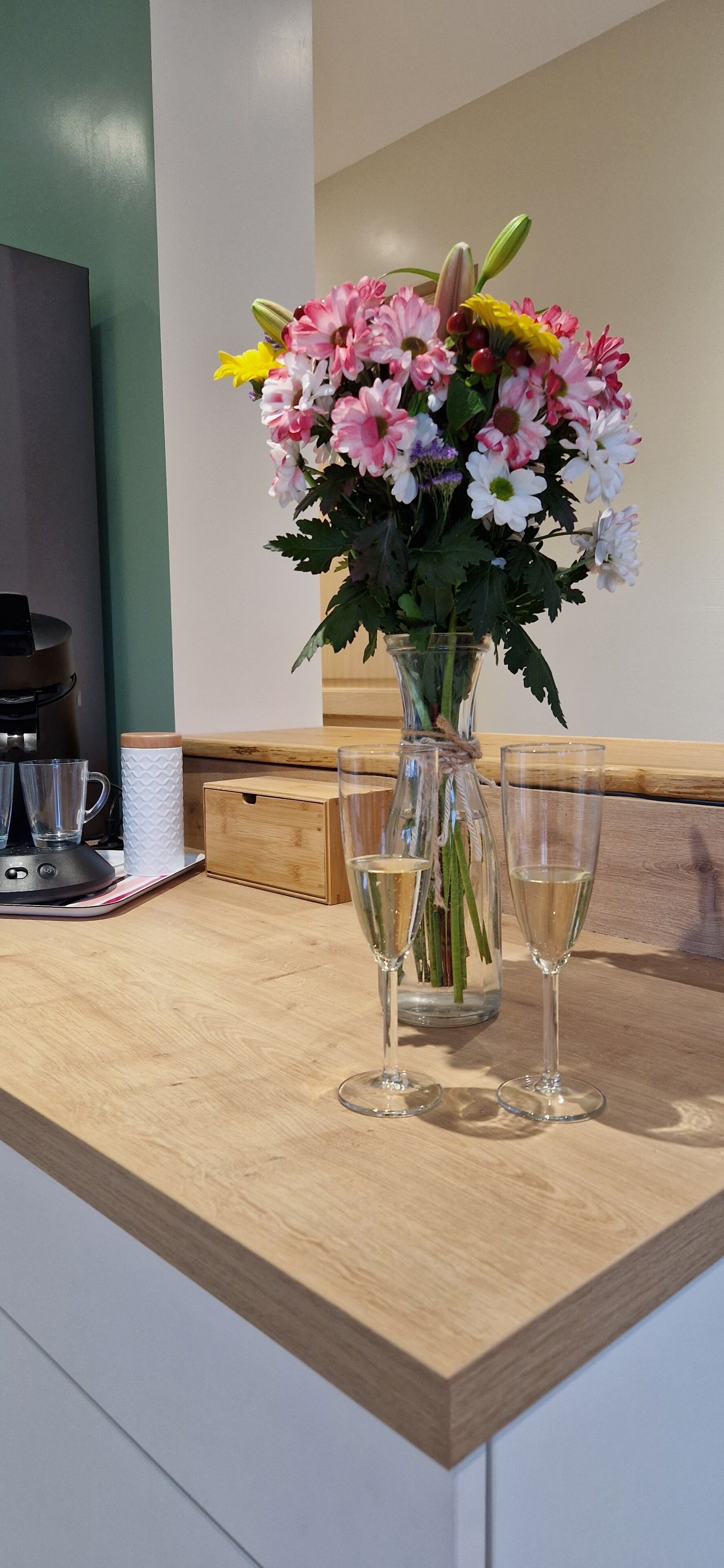 Un comptoir en bois, sur lequel se trouve un vase de fleurs et deux verres contenant un liquide transparent, probablement du vin, ainsi qu’une table avec une machine à café et une boîte.