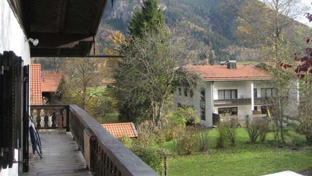 Vista desde un balcón con vistas a un paisaje montañoso, con una casa de techo rojo y otro balcón.