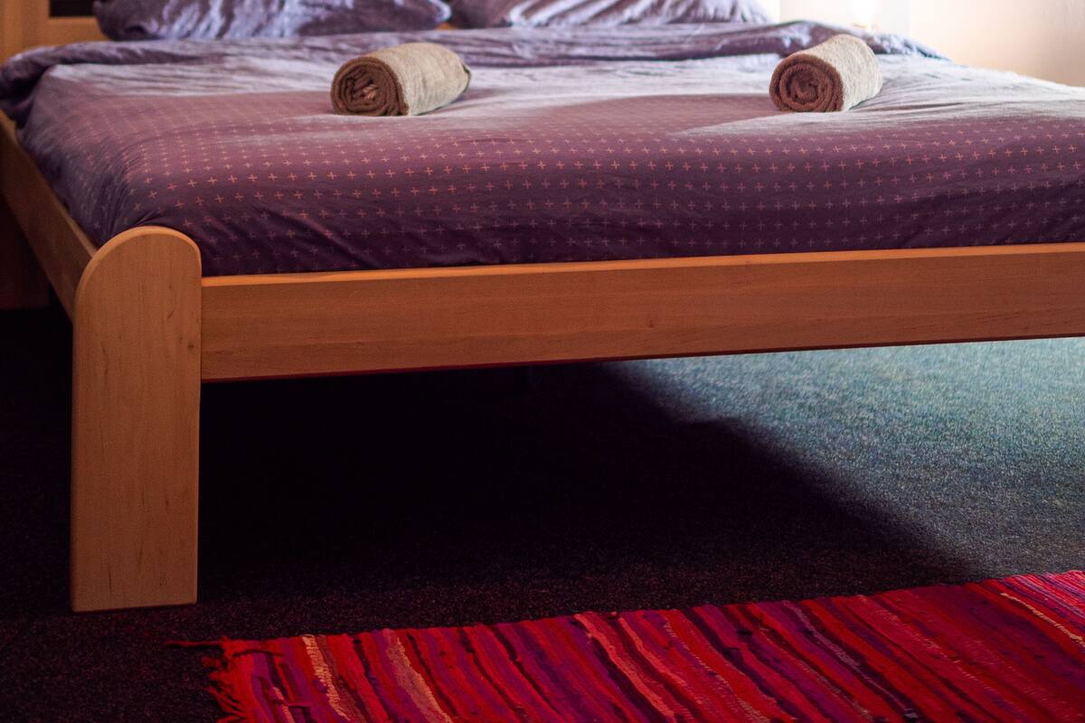 A bedroom with a wooden bed, purple bedding, a red and black rug, and a painting on the wall.