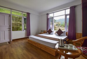 A room with a bed, a wooden bench, a table with cups and a kettle, and large windows with curtains.