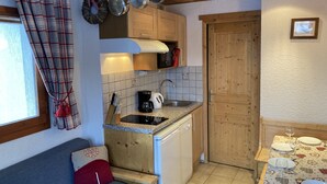 Cocina compacta con estufa integrada, cafetera y microondas. Una puerta de madera conduce a otra habitación, y una mesa de comedor con platos y vasos.