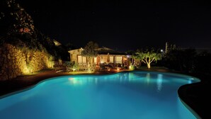 Une piscine la nuit, avec une maison et un espace patio en arrière-plan.