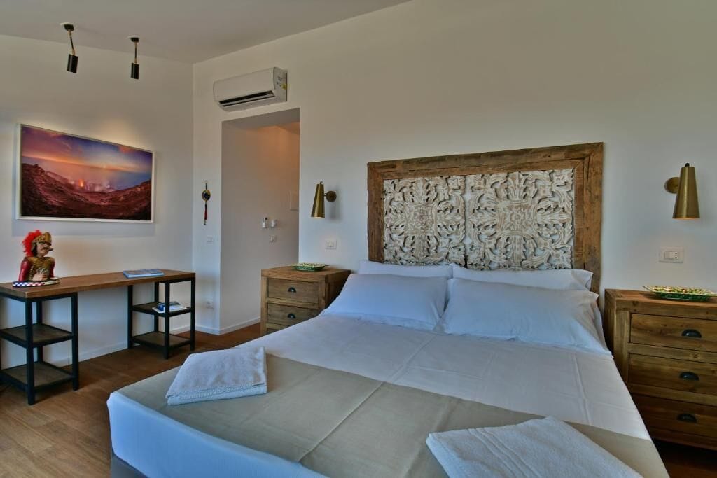 A bedroom with a large bed, a desk, and a chair. There is a painting on the wall and a decorative headboard.
