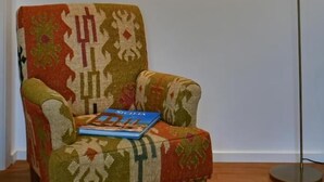 Una silla con un cojín estampado y un libro encima, una lámpara de pie y un cuadro colgado en la pared.