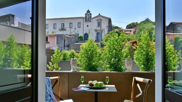 Un balcón con dos sillas de mimbre, una mesita con una fuente de fruta y dos vasos, y vista a un edificio con una torre de campanas.