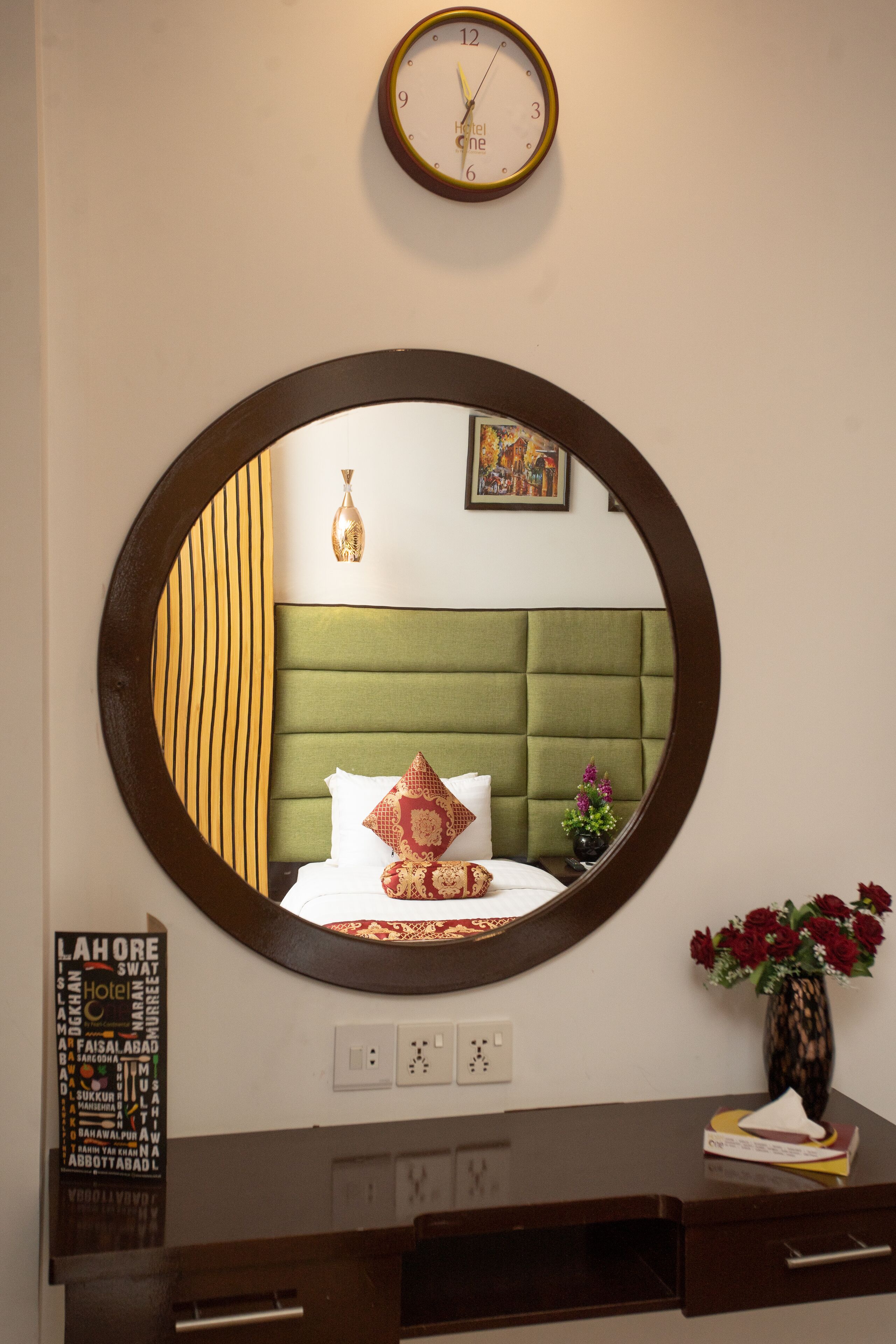 Hotelkamer met ronde spiegel die een bed met groene kapton, een klok en een bloevas weerkaatst.