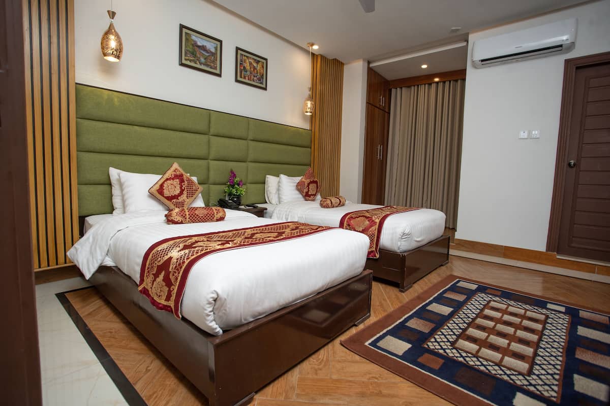 Una habitación de hotel con dos camas, un cabecero tapizado en verde, una alfombra estampada y un equipo de aire acondicionado.