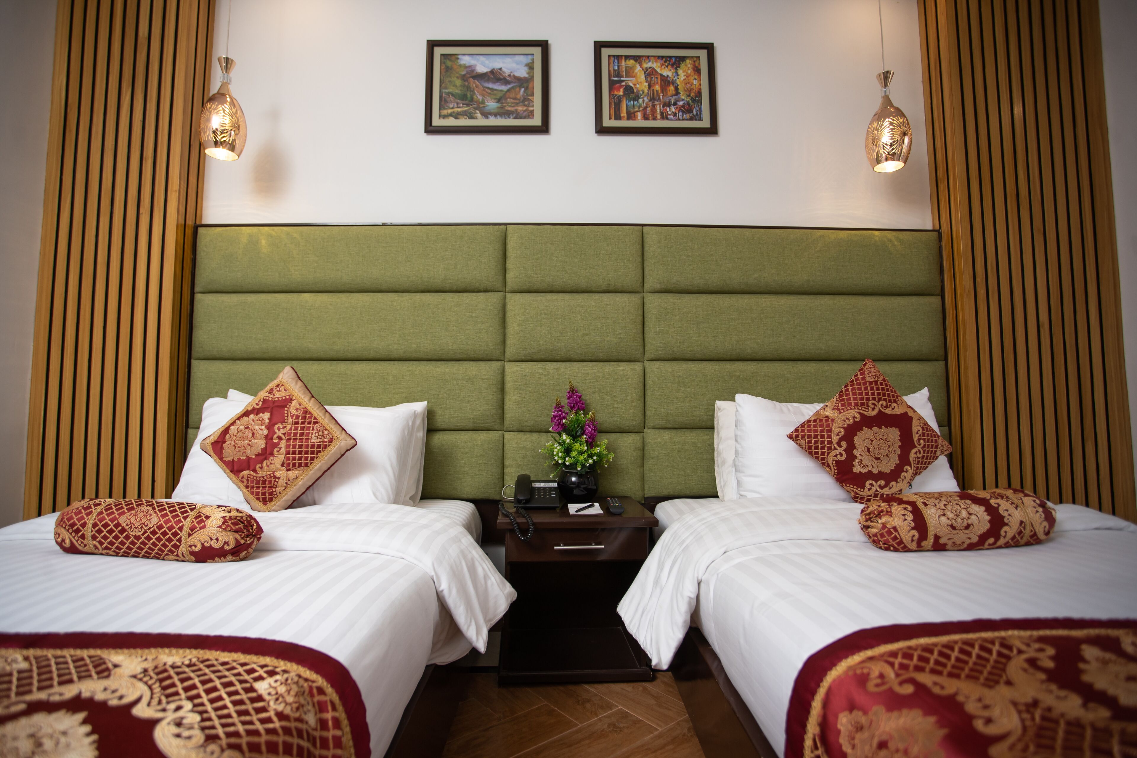 Habitación de hotel con dos camas, cada una con almohada de patrón rojo y dorado, cabecera tapizada en verde y una mesita de noche con un teléfono y un florero.