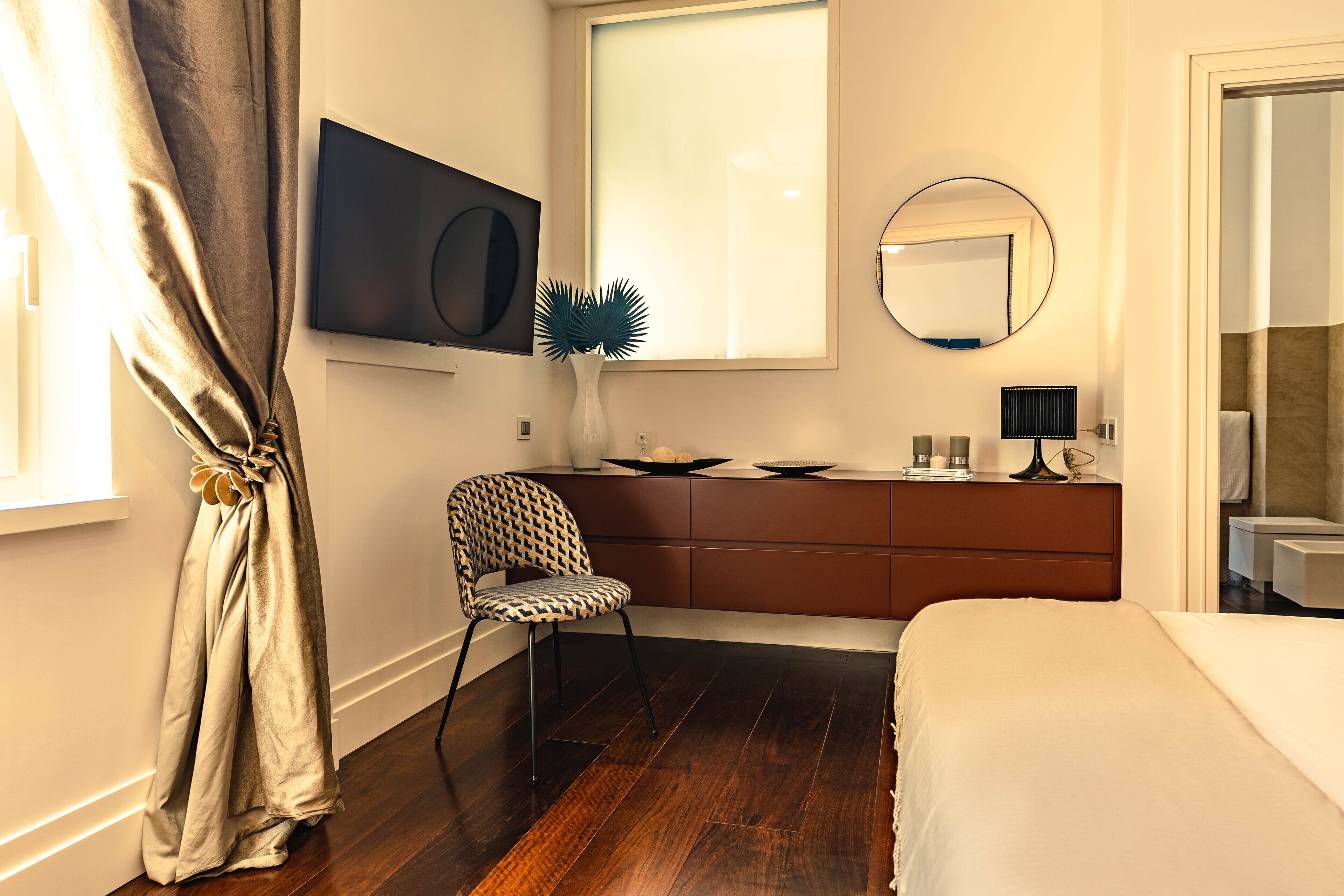 Une chambre à coucher moderne avec un plancher en bois, un lit, une commode, une chaise, un téléviseur et une fenêtre avec des rideaux.