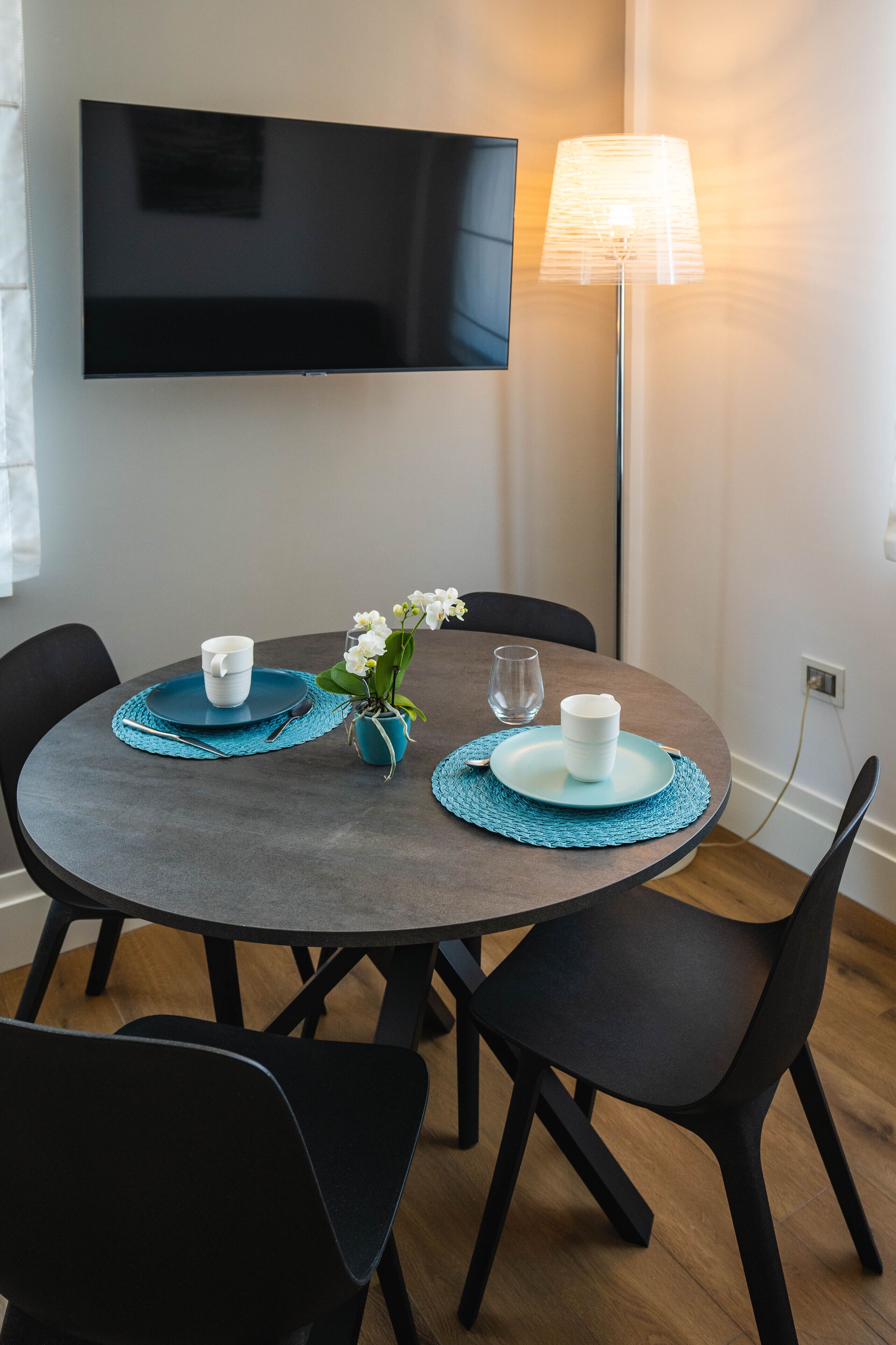 Een ronde eettafel met een flatscreen televisie aan de muur erboven, een vloerlamp en een vaas met witte bloemen.