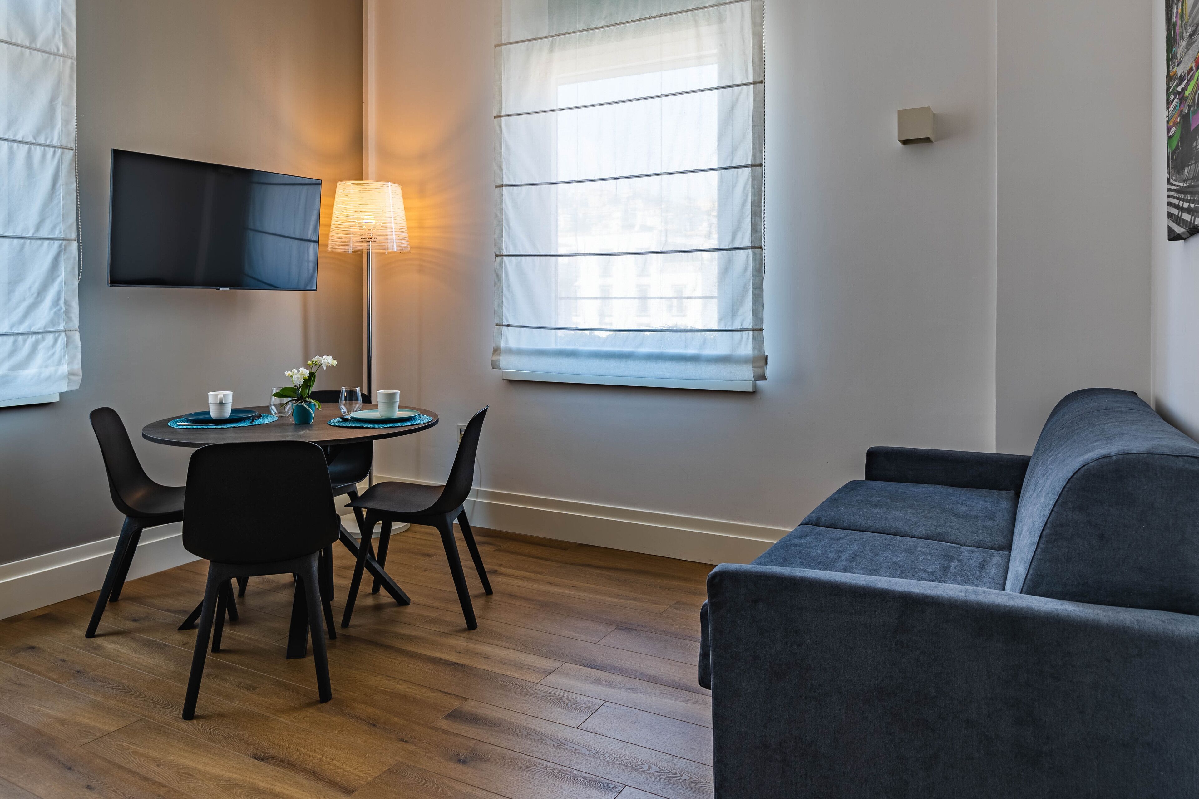 Ett modernt vardagsrum med en grå soffa, ett runt matbord med stolar, en platt-TV som är monterad på väggen och en golvlampa.