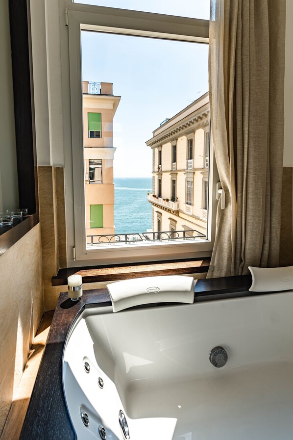 A bathroom with a bathtub, a window with a view of the sea and buildings, and a curtain partially drawn. - AD MAIORA Boutique Suite (Naples)