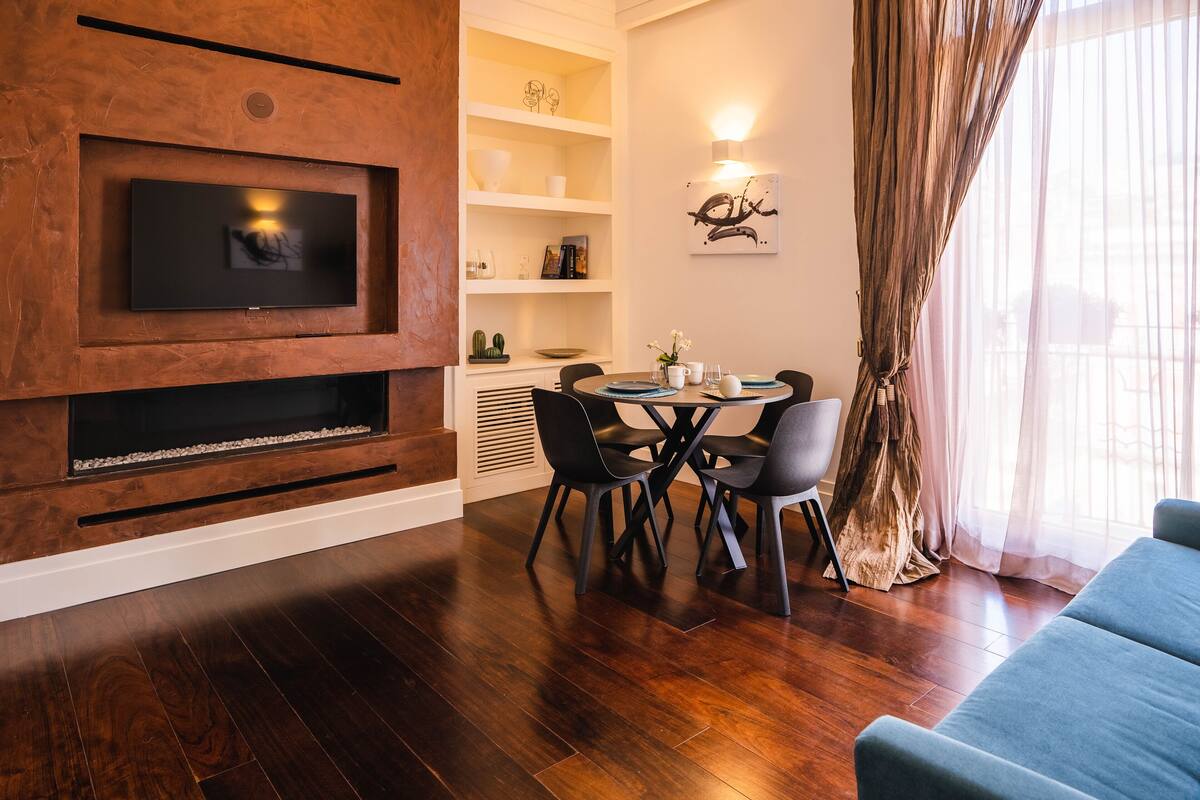A modern living room with a flat-screen TV mounted on a wooden wall, a dining area with a table and chairs, and a large window with sheer curtains.