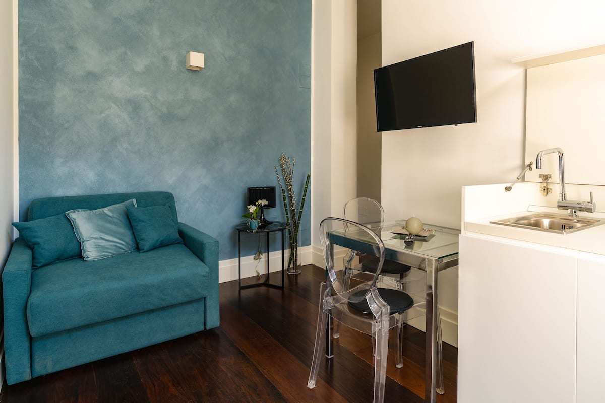 A compact living space with a teal sofa, a clear glass table, and a modern kitchen area with a sink and a mounted TV.
