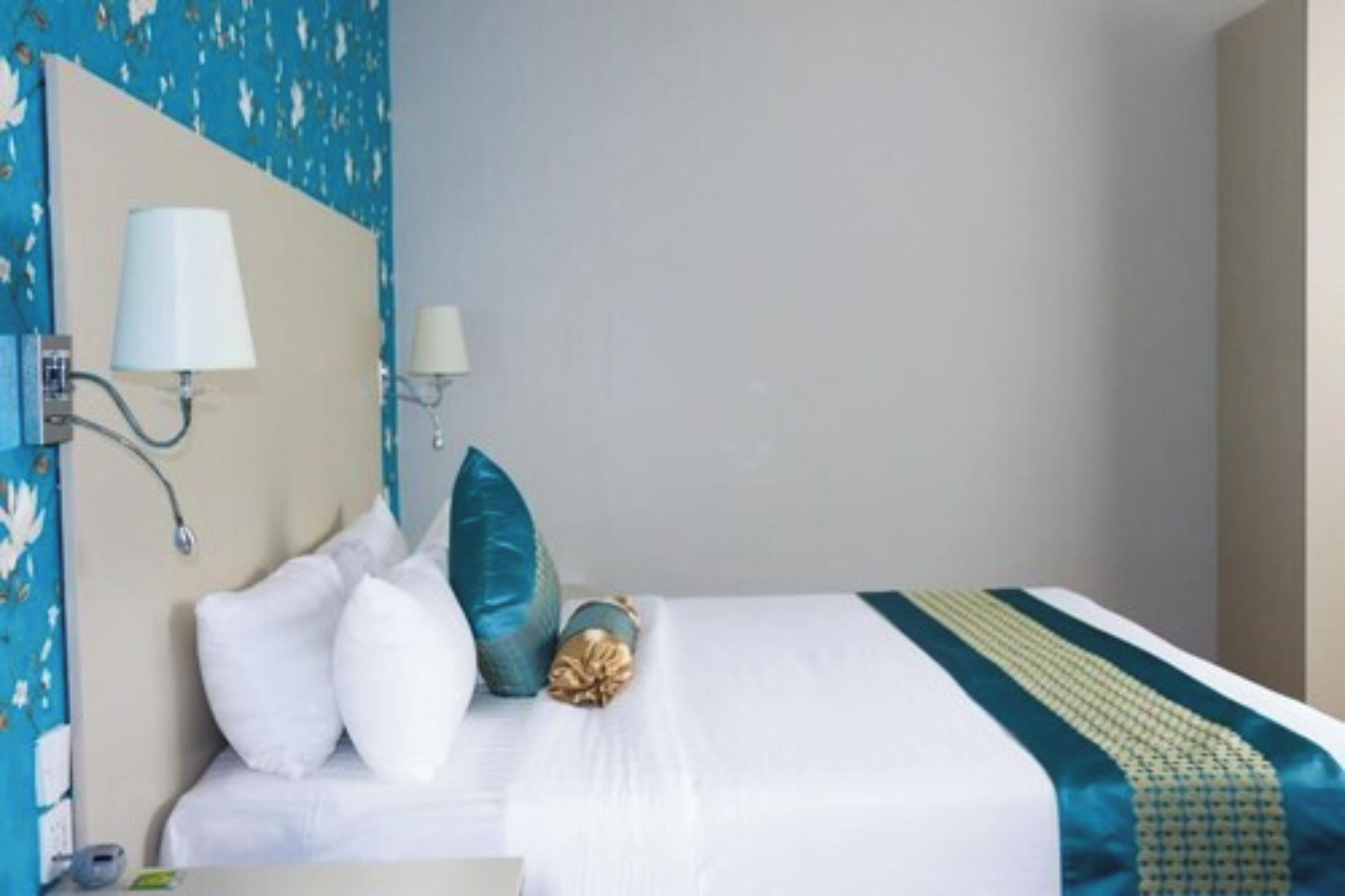 Een hotelkamer met een bed, wit beddengoed, een blauwgroene en gouden plaid, en een wandlamp.