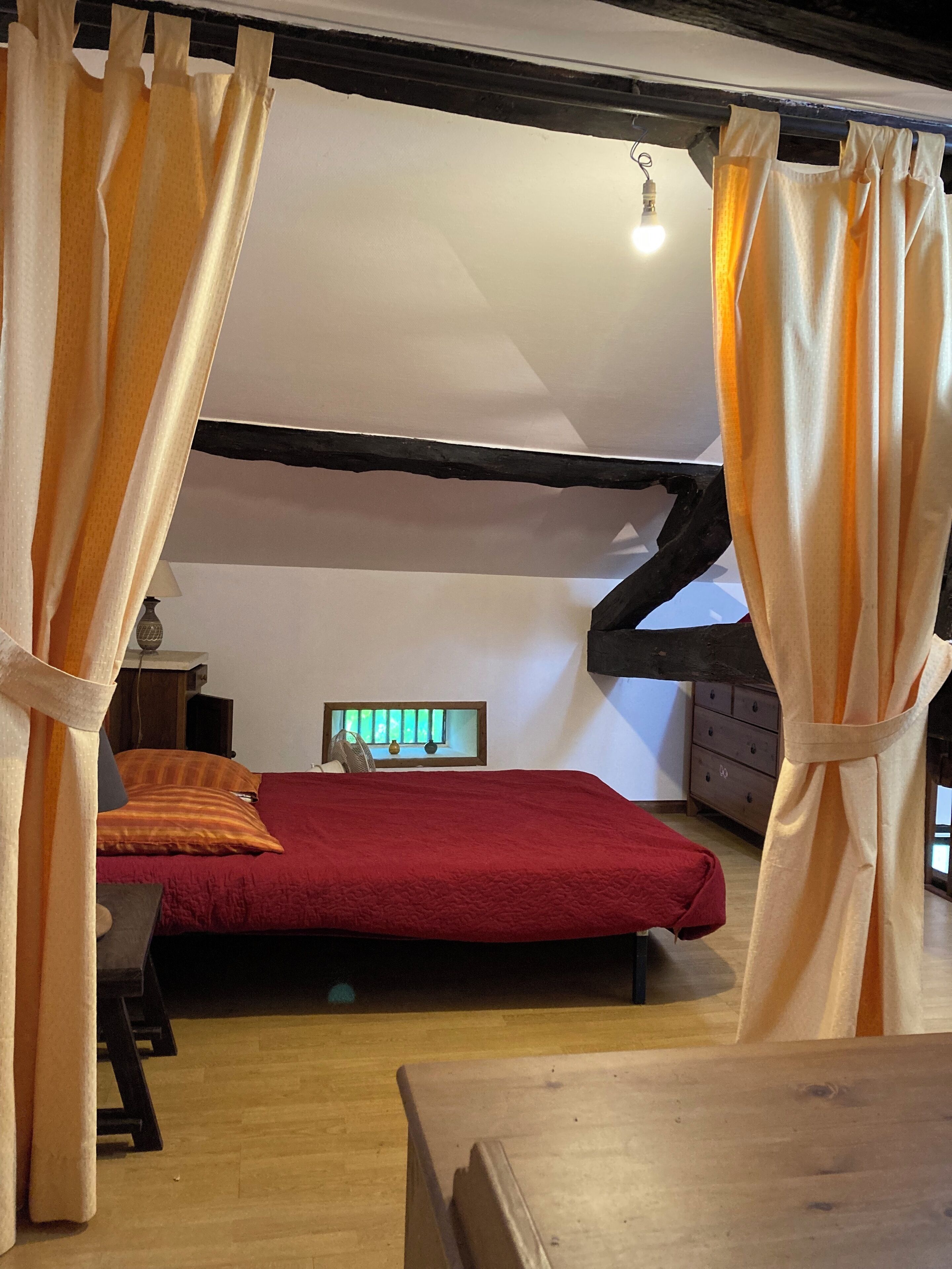 Un dormitorio con techo de madera, una cama con colcha roja, una mesita y una ventana con vistas al verdor.