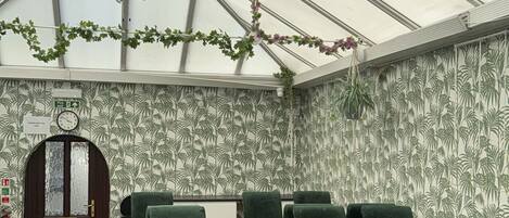 Een kas-achtig vertrek met een hoge plafond, groene stoelen en een houten deur.
