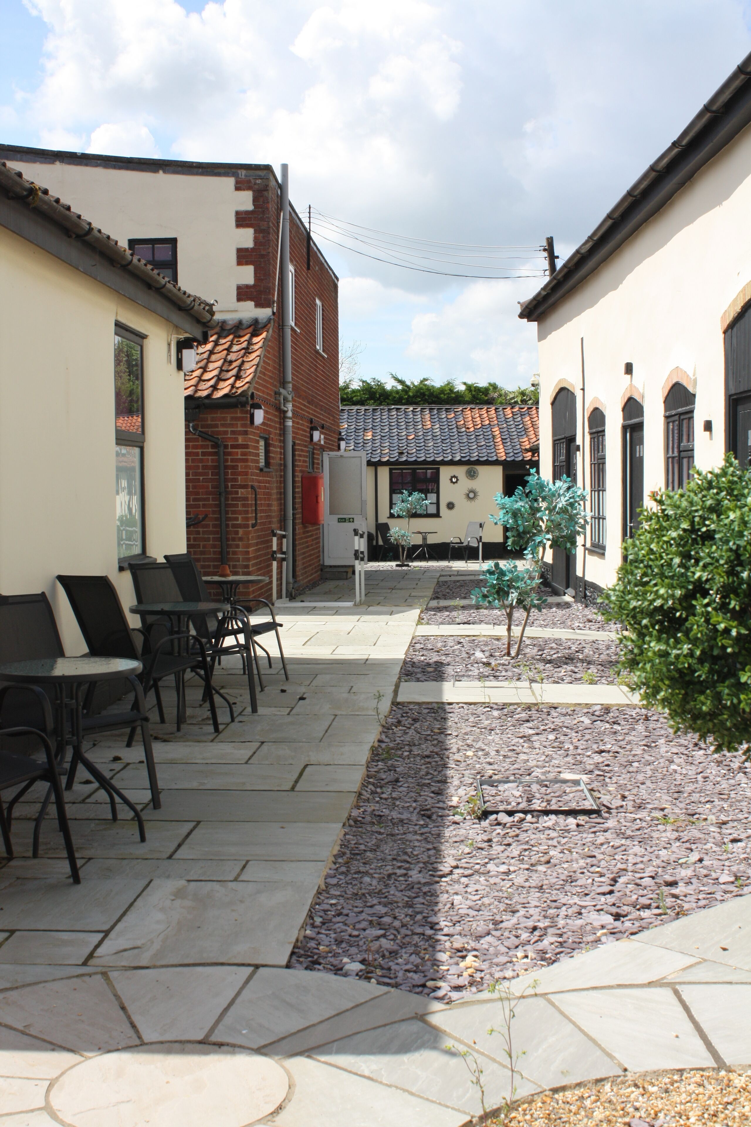 An outdoor patio area with stone flooring, metal chairs, and tables. There are buildings with brick and stucco exteriors, and a small tree in the center.