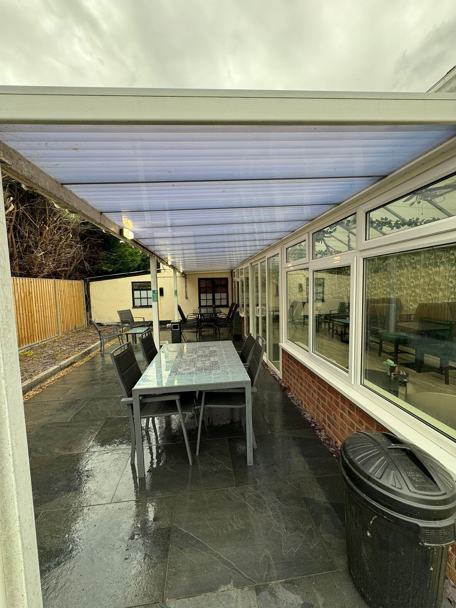 A covered patio area with a table and chairs, a trash bin, and a view of the outside through large windows.