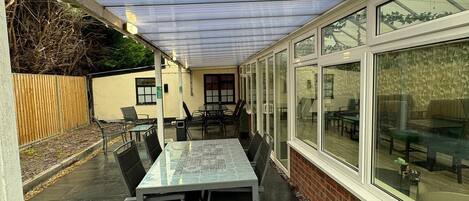 A covered patio area with a table and chairs, a trash bin, and a view of the outside through large windows.
