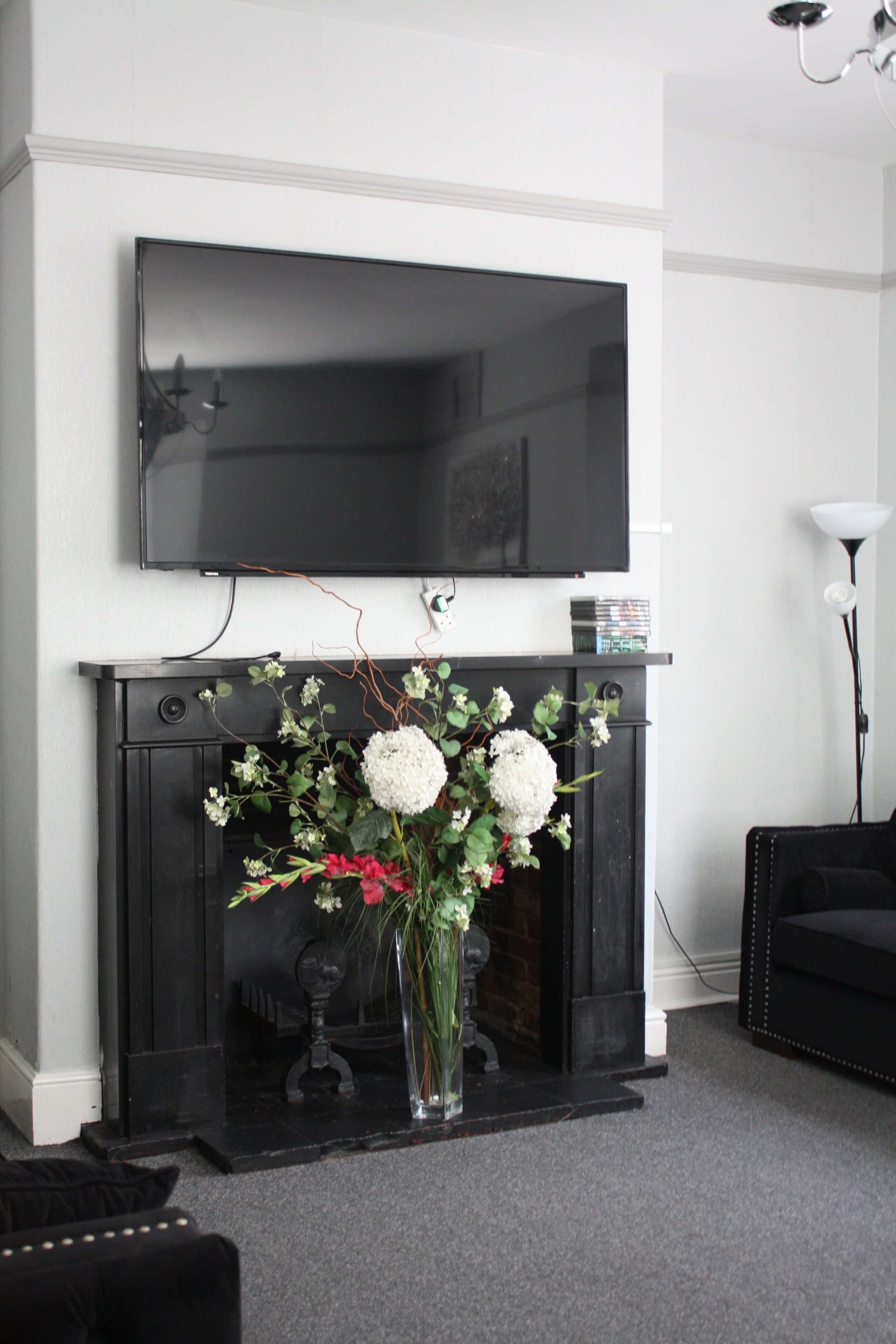 Ein Wohnzimmer mit Flachbildfernseher über einem Kamin, einer Vase mit Blumen auf dem Kaminvorsprung und einem schwarzen Sofa.