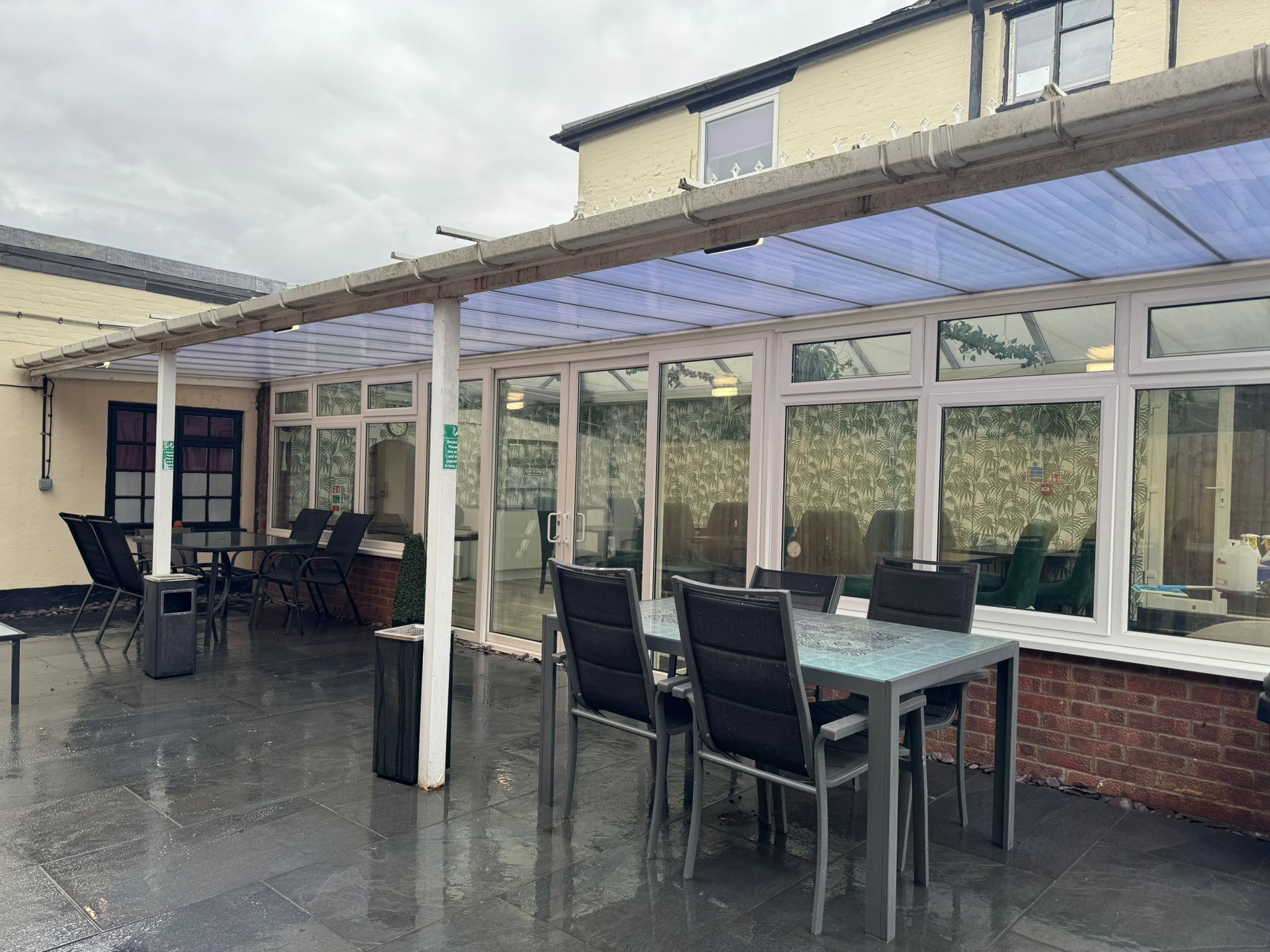 An outdoor patio area with tables and chairs, a covered section with a glass roof, and a brick wall.