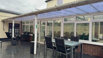 Un espace extérieur aménagé en terrasse, avec des tables et des chaises, une partie couverte dotée d’un toit en verre et un mur en briques.